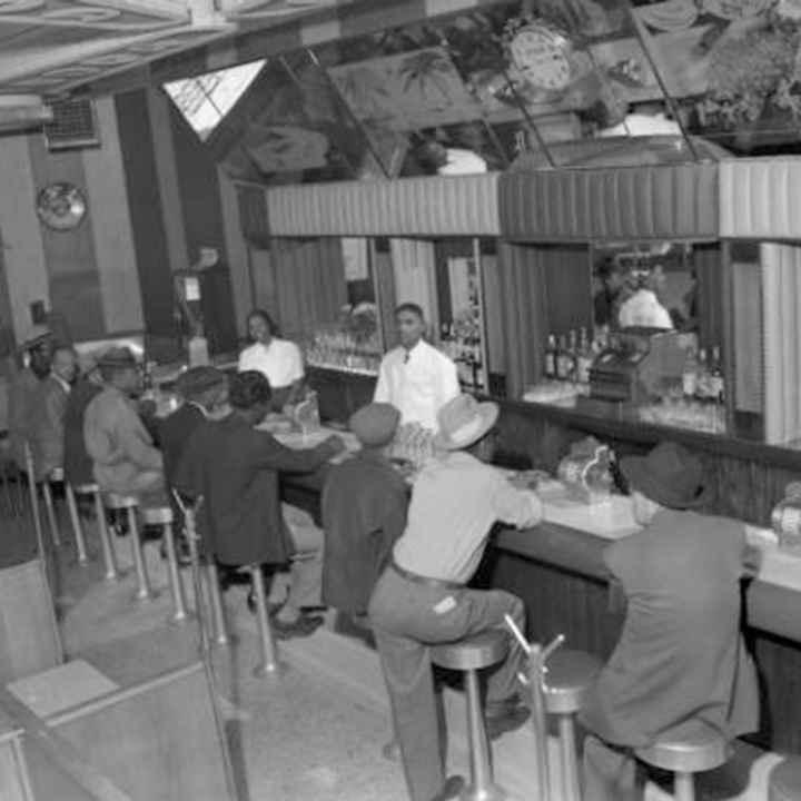 一名男性(左)和一名女性调酒师在715俱乐部的室内照片，该俱乐部位于Five Points社区的东26(26)大道715号. 一排男性顾客坐满了酒吧的座位. 他们戴着软呢帽和报童帽.